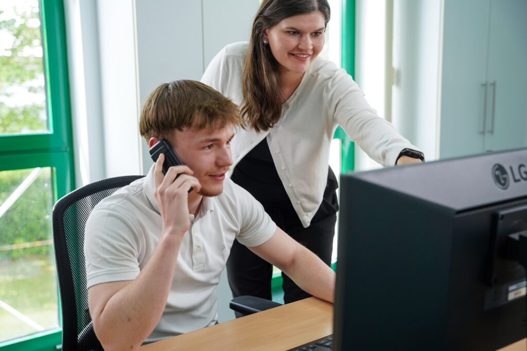 Global & Service Team Global & Service Team, eine Mitarbeiterin und ein Mitarbeiter schauen auf den PC, der Mitarbeiter telefoniert, die Mitarbeiterin zeigt auf den PC